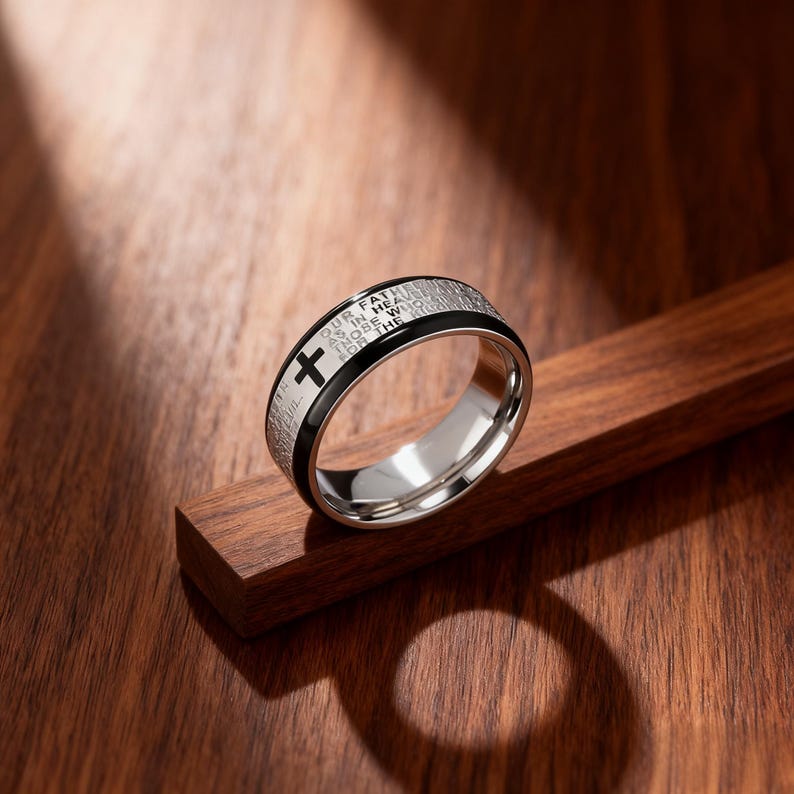 K&ouml;nnte beinhalten: Ein Edelstahlring mit schwarzem Band und Kreuzdesign. Der Ring hat eingravierten Text am &auml;u&szlig;eren Rand. Der Ring wird auf einer Holzoberfl&auml;che pr&auml;sentiert.