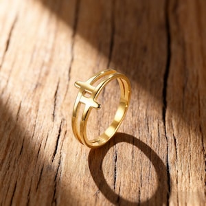 Peut inclure: Une bague dorée avec un double anneau et un motif en forme de croix stylisée. La bague, en métal brillant, est posée sur une surface en bois. L'ombre de la bague est visible sur le bois.