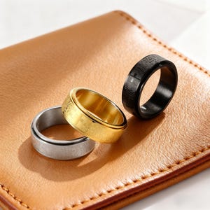 May include: Three metal rings in silver, gold, and black, each with a spinning center band. The rings have engraved text and a cross design. The rings are displayed on a brown leather surface.