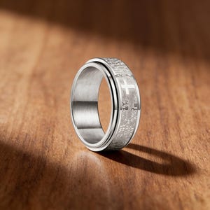 K&ouml;nnte beinhalten: Ein silberfarbener Ring mit Kreuzdesign und graviertem Text. Der Ring hat ein drehbares Au&szlig;enband. Das Kreuz ist ein markantes Merkmal auf der Oberfl&auml;che des Rings. Pr&auml;sentiert auf einer Holzoberfl&auml;che.