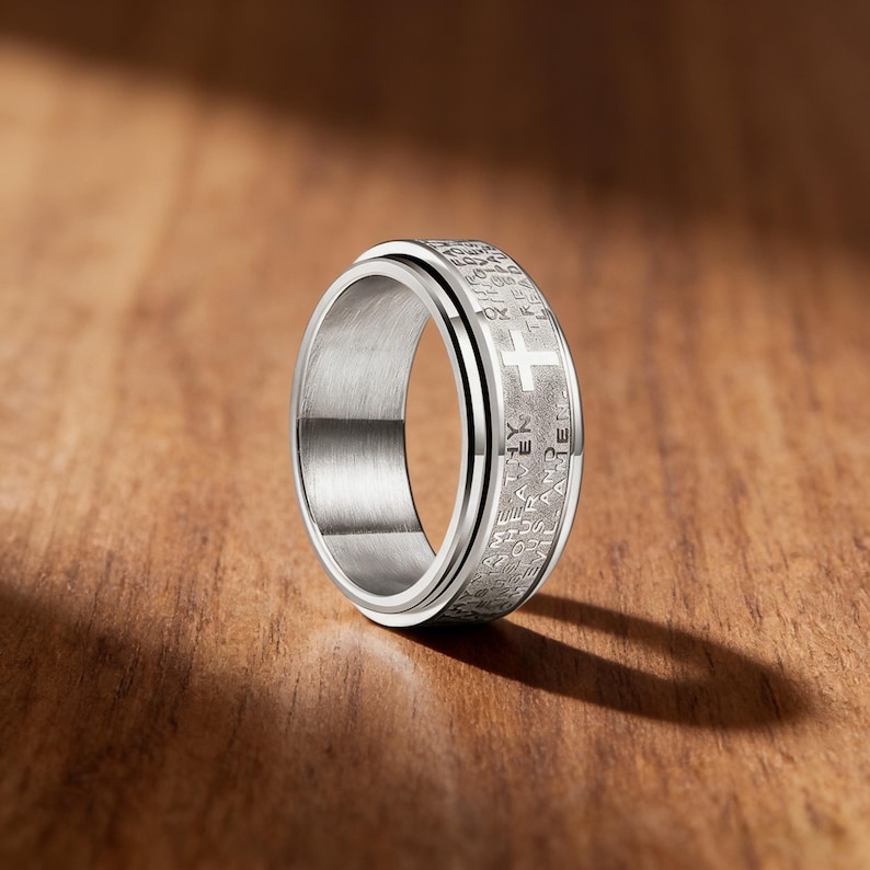 K&ouml;nnte beinhalten: Ein silberfarbener Ring mit Kreuzdesign und graviertem Text. Der Ring hat ein drehbares Au&szlig;enband. Das Kreuz ist ein markantes Merkmal auf der Oberfl&auml;che des Rings. Pr&auml;sentiert auf einer Holzoberfl&auml;che.