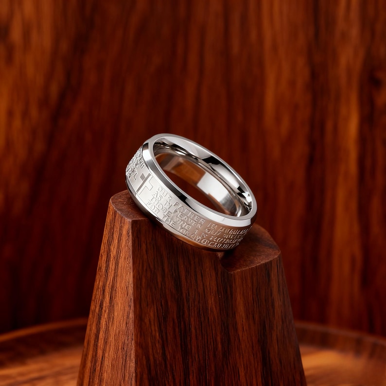 K&ouml;nnte beinhalten: Ein silberfarbener Ring mit Kreuzmuster und eingraviertem Text. Der Ring wird auf einem Holzst&auml;nder vor einem dunkelbraunen Hintergrund pr&auml;sentiert. Der Text auf dem Ring ist teilweise sichtbar.