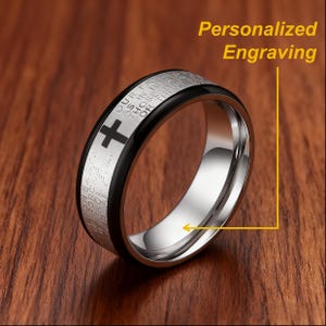 K&ouml;nnte beinhalten: Ein Edelstahlring mit schwarzem Band und Kreuzdesign. Der Ring hat eingravierten Text und die Worte "Personalized Engraving". Der Ring wird auf einer Holzoberfl&auml;che pr&auml;sentiert.