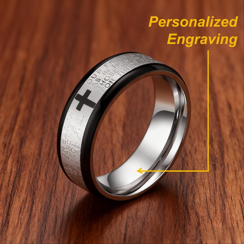 K&ouml;nnte beinhalten: Ein Edelstahlring mit schwarzem Band und Kreuzdesign. Der Ring hat eingravierten Text und die Worte "Personalized Engraving". Der Ring wird auf einer Holzoberfl&auml;che pr&auml;sentiert.