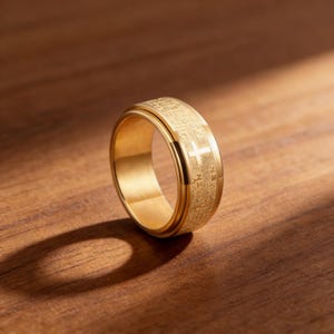 K&ouml;nnte beinhalten: Ein goldfarbener Ring mit Kreuz und graviertem Text. Der Ring aus gl&auml;nzendem Metall liegt auf einer Holzoberfl&auml;che. Das Kreuz befindet sich mittig auf dem Band. Ein religi&ouml;ses Accessoire f&uuml;r den t&auml;glichen Gebrauch.