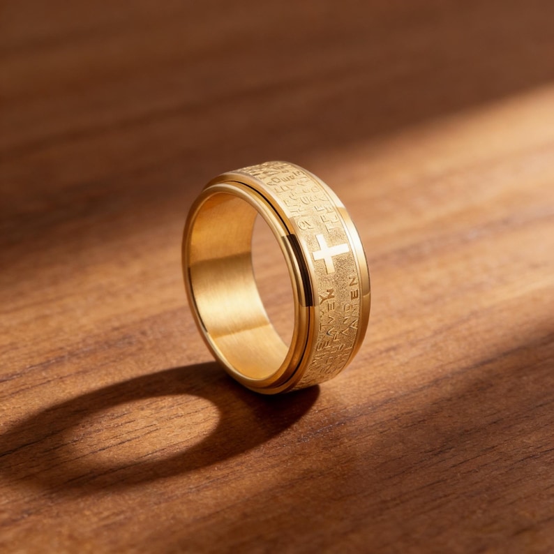 K&ouml;nnte beinhalten: Ein goldfarbener Ring mit Kreuz und graviertem Text. Der Ring aus gl&auml;nzendem Metall liegt auf einer Holzoberfl&auml;che. Das Kreuz befindet sich mittig auf dem Band. Ein religi&ouml;ses Accessoire f&uuml;r den t&auml;glichen Gebrauch.