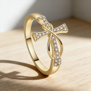 Peut inclure: Bague dorée ornée d'une croix incrustée de petits diamants transparents. La croix est entrelacée avec un symbole infini, créant un design unique et élégant. La bague est posée sur une surface en bois clair.