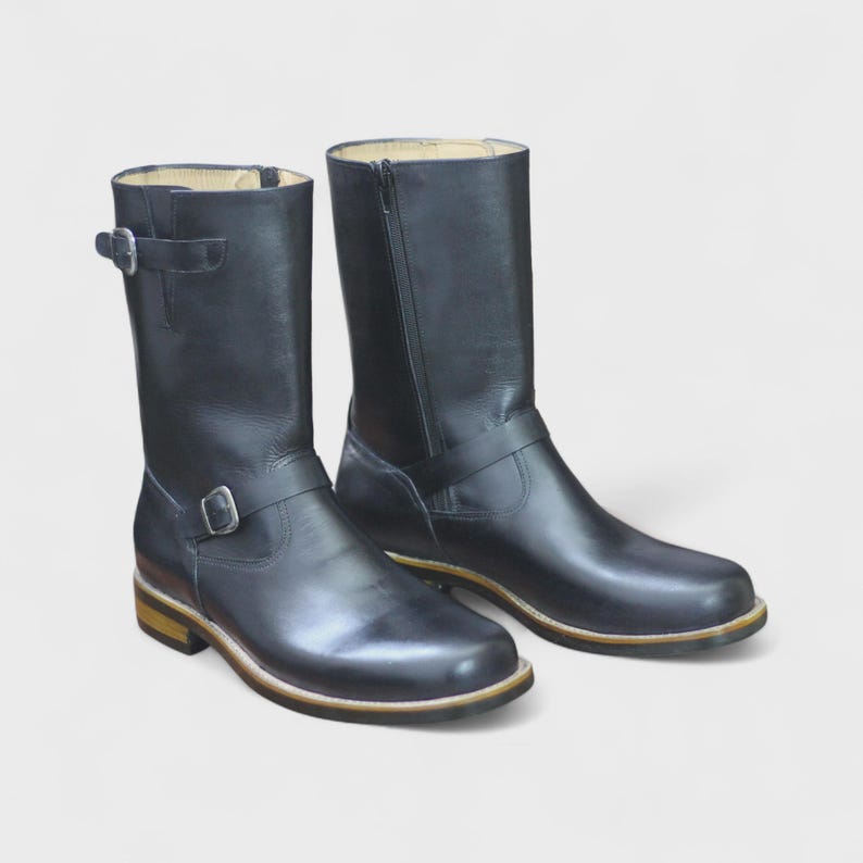 May include: A pair of black leather motorcycle boots. Each boot has a buckle strap near the ankle and a zip on the inner side. The soles have a light brown trim.