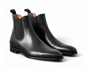 Pode incluir: Um par de botas Chelsea de couro preto. As botas têm uma aparência elegante e polida, com painéis laterais elásticos e abas de puxar. As solas são escuras e as botas estão em um fundo branco.