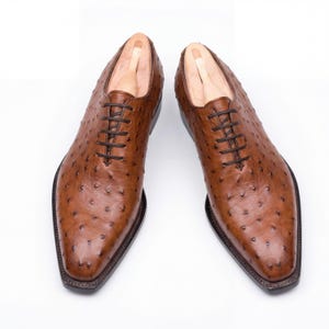 Men wearing handmade cognac full quill ostrich wholecut Oxford shoes with charcoal grey tailored dress trousers on modern concrete surface in architectural setting