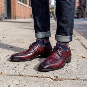 Peut inclure: Chaussures de ville en cuir bordeaux avec un motif peau de crocodile. Les chaussures ont des lacets et des semelles noirs. La personne porte des chaussettes à motifs et un jean bleu foncé avec les revers retroussés.