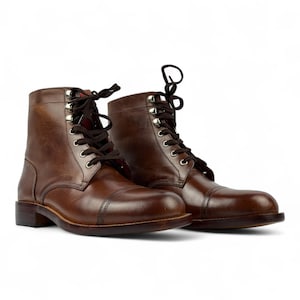 May include: A pair of brown leather boots with a cap toe design. The boots have dark brown laces, metal eyelets, and a low heel. The leather has a polished finish, and the boots are set against a white background.