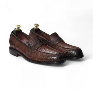 May include: A pair of brown leather loafers with a textured snakeskin pattern. The shoes have a classic penny loafer design with a rounded toe and a low heel. Shoe trees are inserted inside the shoes.