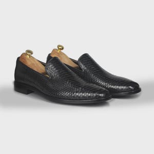 May include: A pair of black leather loafers with a textured, scale-like pattern. The shoes have a low heel and are displayed with wooden shoe trees. The loafers are a classic style.