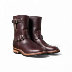 Könnte beinhalten: Ein Paar dunkelbraune Leder-Ankle Boots. Die Stiefel haben Schnallenriemen um den Knöchel und oben am Stiefel. Die Sohlen haben ein geschichtetes Design mit hellbraunen und schwarzen Zierleisten. Die Stiefel sind vor weißem Hintergrund.