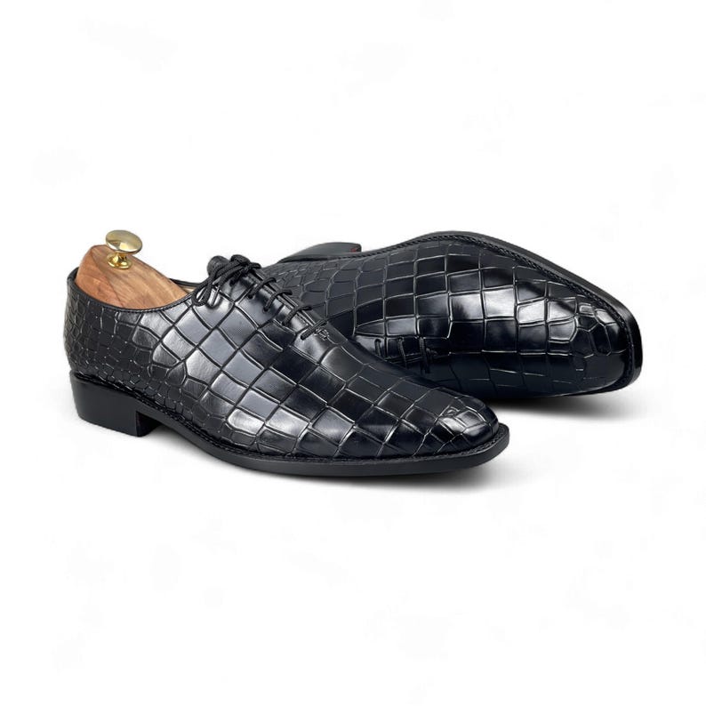 Op de afbeelding: Zwarte leren herenschoenen met een textuur, krokodillenpatroon. De schoenen zijn geregen en hebben een lage hak. E&eacute;n schoen staat rechtop en de andere ligt op zijn kant. Een houten schoenspanner zit in de rechtopstaande schoen.