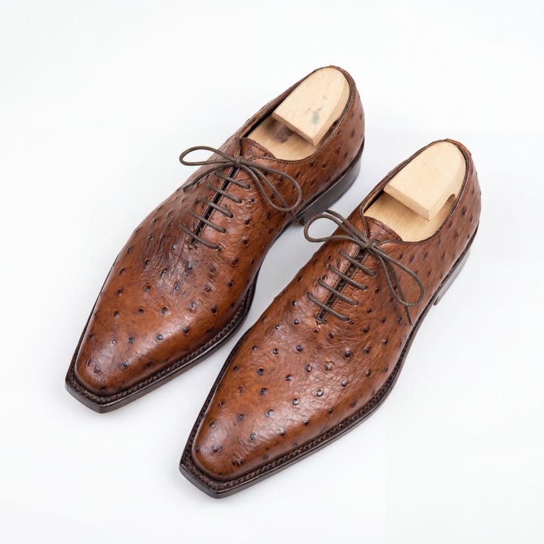 Pair of men handmade full quill ostrich leather wholecut Oxford shoes in cognac tan displayed on wooden podium inside luxury leather goods boutique with warm ambient lighting