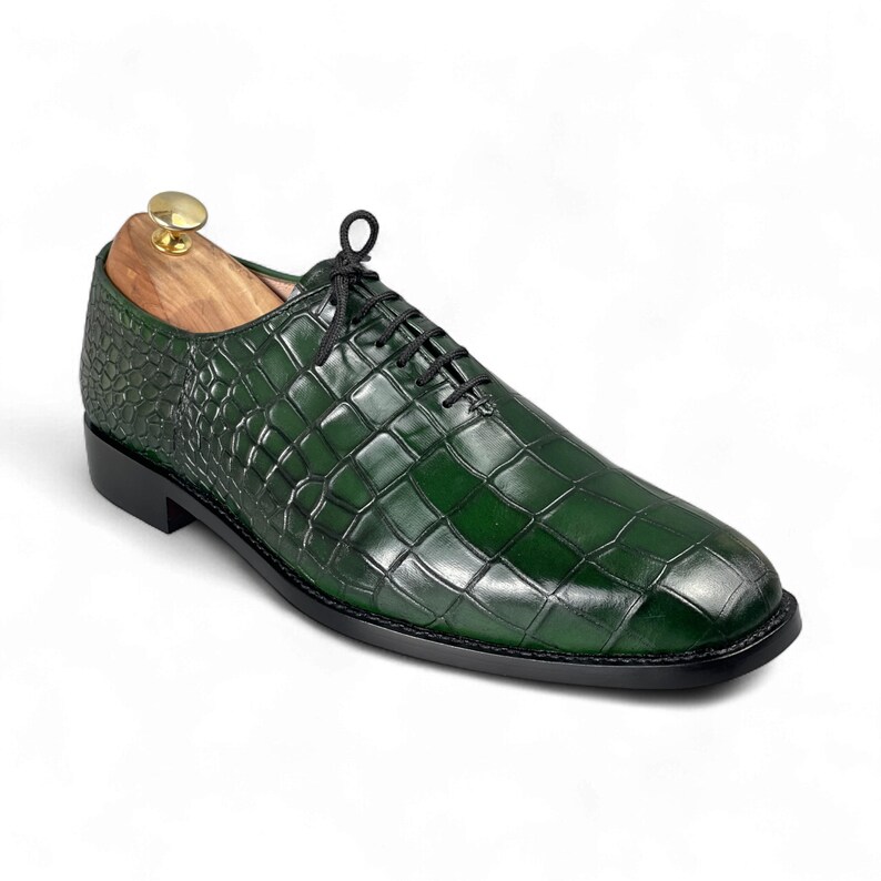 Op de afbeelding: Een enkele groene leren herenschoen met een krokodillenpatroon. De schoen heeft zwarte veters en een zwarte zool. Een houten schoenspanner met een gouden knop zit erin.