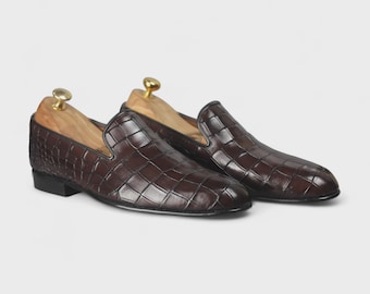 May include: A pair of brown leather loafers with a crocodile skin pattern. The shoes have a classic slip-on design with a low heel and are displayed with wooden shoe trees.