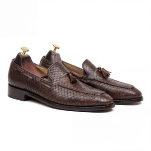 May include: A pair of brown leather loafers with a textured, scale-like pattern. Each shoe features a small tassel and is displayed with a wooden shoe tree inside. The shoes have a classic, slip-on design with a low heel.