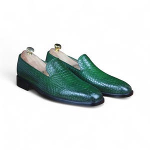 May include: A pair of green leather loafers with a textured, reptile-like pattern. The shoes have a classic slip-on design with a low heel and are displayed with wooden shoe trees.