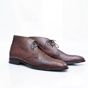 May include: A pair of brown ankle boots featuring a textured, scale-like pattern across the exterior. These boots have a rounded toe, a low heel, and thin dark brown laces threaded through two eyelets on each side of the upper ankle section.