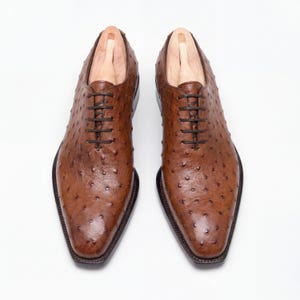 Street style shot of men wearing cognac ostrich wholecut Oxford shoes with rolled selvedge dark denim jeans on urban cobblestone street with pedestrians in background