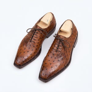 Men wearing cognac full quill ostrich wholecut Oxford shoes with black trousers crossed legs on rustic wood floor inside vintage cafe holding coffee cup