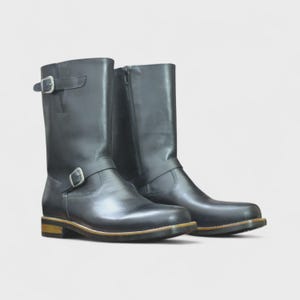 May include: A pair of black leather boots with a mid-calf height. Each boot features a buckle strap and a side zip. The soles have a light brown trim. The boots are designed for durability and style.