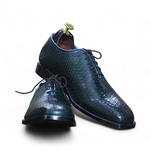 Könnte beinhalten: Ein Paar dunkelgrüne Lederschuhe mit strukturiertem Muster. Die Schuhe haben schwarze Sohlen, türkisfarbene Schnürsenkel und einen hölzernen Schuhspanner in einem Schuh. Der Hintergrund ist weiß.