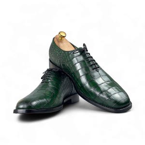Op de afbeelding: Een paar groene leren herenschoenen met een krokodillenleerpatroon. De schoenen hebben zwarte veters en een zwarte zool. Een houten schoenspanner is in &eacute;&eacute;n schoen geplaatst. Op een witte achtergrond.