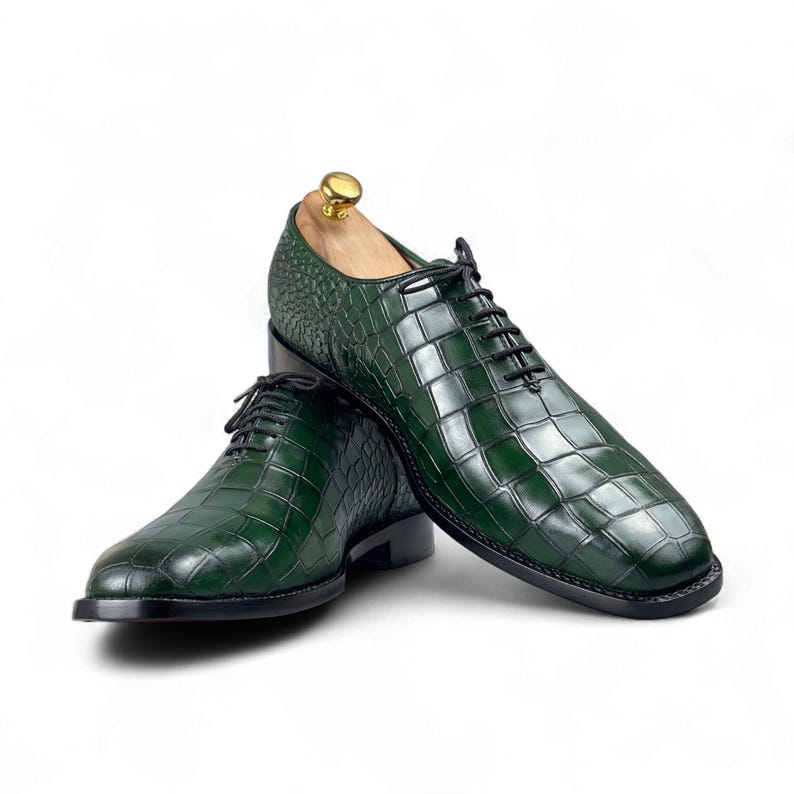 Op de afbeelding: Een paar groene leren herenschoenen met een krokodillenleerpatroon. De schoenen hebben zwarte veters en een zwarte zool. Een houten schoenspanner is in &eacute;&eacute;n schoen geplaatst. Op een witte achtergrond.