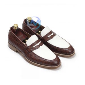 May include: A pair of brown and white leather loafers. The shoes have a textured brown leather upper with a white vamp and strap. The soles are a light brown color. The shoes are displayed on a white background.