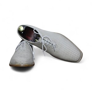 Könnte beinhalten: Ein Paar hellgraue Lederschuhe mit strukturierter, schuppenartiger Musterung und weißen Schnürsenkeln. Die Schuhe haben eine quadratische Spitze. Ein Schuh steht aufrecht, der andere ist abgewinkelt und zeigt das Innere. Elegante Schuhe.