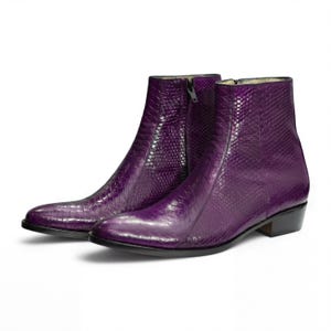 Puede incluir: Un par de botines morados con un patrón texturizado similar a la piel de serpiente. Los botines tienen punta afilada, cremallera lateral y tacón bajo. Los botines son de material brillante y están sobre fondo blanco.