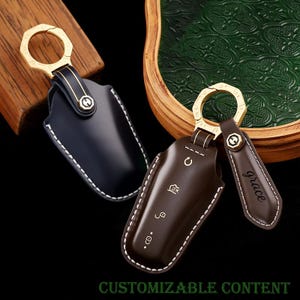 May include: Two leather key fob covers, one black and one brown, with gold-coloured key rings. The brown cover has white stitching and embossed icons. The word "Grace" is on a brown leather tag. The text "CUSTOMIZABLE CONTENT" is at the bottom.