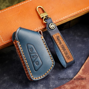 Pode incluir: Uma capa de chave de carro em couro azul e um porta-chaves a condizer. A capa da chave tem costuras laranja e botões em relevo. O porta-chaves tem uma etiqueta de couro com o texto "Name. 1995.06.01". As palavras "Customizable content" também são visíveis.
