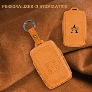 Könnte beinhalten: Orangenes Leder-Schlüsselanhänger-Etui mit silbernem Schlüsselring. Das Etui hat einen genähten Rand und geprägte Symbole. Der Text "PERSONALISIERTE ANPASSUNG" steht oben. Ein zweites Etui mit dem Namen "ALEPPI" ist ebenfalls abgebildet.
