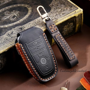 May include: Black leather key fob cover with orange stitching and a matching key strap. The key fob cover has embossed buttons and a key symbol. The key strap is personalized with the name "David" and the date "1995.06.01".