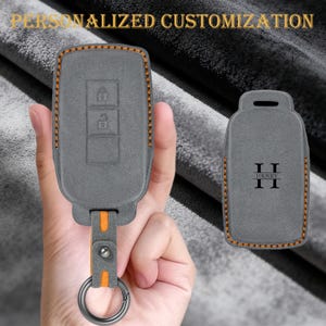 Może przedstawiać: Szara osłona na brelok z pomarańczowymi przeszyciami i pasującym brelokiem. Osłona ma trzy przyciski i skórzany pasek z metalowym pierścieniem. Tekst "SPERSONALIZOWANE DOSTOSOWANIE" znajduje się u góry obrazu.