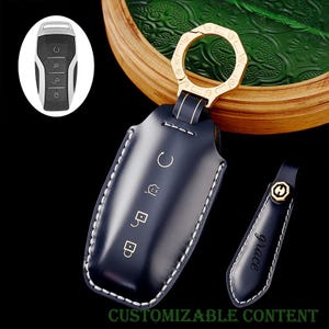 Puede incluir: Un estuche para llavero de cuero azul marino con costuras blancas y herrajes dorados. El estuche tiene símbolos en relieve y una etiqueta de llave a juego con la palabra "grace". También se muestra un mando a distancia para la llave del coche.