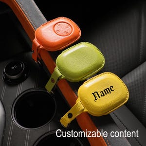 May include: Three leather car key fob covers in orange, lime green, and yellow. Each cover features a strap with a metal snap button. The yellow cover displays the word Name in a black gothic font. The covers are textured and rectangular.