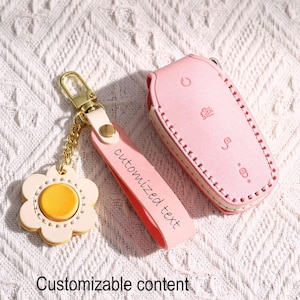 Puede incluir: Un estuche para llavero de cuero rosa y un juego de llavero a juego. La funda del llavero tiene símbolos de botones y un borde cosido. El llavero presenta un dije de flor con una cadena y herrajes dorados. La correa dice "customized text".