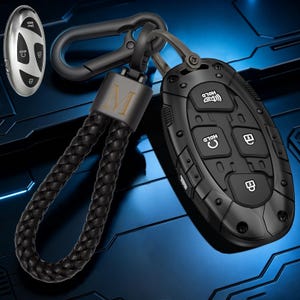 Könnte beinhalten: Eine schwarze Autoschlüsselhülle mit fünf Tasten, die Symbole für Verriegeln, Entriegeln und Halten zeigen, befestigt an einem schwarzen geflochtenen Lederband und einem Metallkarabiner. Ein silberner Schlüssel mit schwarzen Tasten ist oben links.