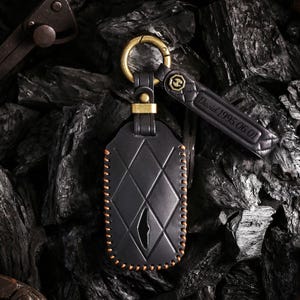 May include: Black leather key fob with a diamond pattern and orange stitching. The fob has a gold-coloured clasp and a matching leather tag with the engraved text "David 1995.06.01". The key fob is designed to hold a car key.