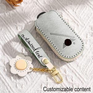 Può includere: Un portachiavi in pelle grigio chiaro e un portachiavi abbinato con un ciondolo a forma di fiore. Il portachiavi ha un bottone nero e un design a mazza da golf. La cinghia del portachiavi dice "testo personalizzato".