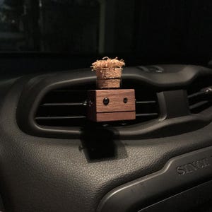 May include: A wooden car air vent clip with a small, decorative plant on top. The clip is a dark brown, rectangular shape with two black screws and two black dots that resemble eyes. The plant is in a small, woven basket.