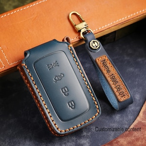 Può includere: Una custodia in pelle blu scuro per chiave auto con cuciture arancioni e un cinturino portachiavi in pelle abbinato. Il cinturino presenta un'etichetta rettangolare in pelle con il testo in rilievo Name.1995.06.01 e un moschettone in metallo color ottone.