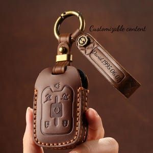 Puede incluir: Funda de llavero de cuero marrón con un cierre dorado y una correa a juego. La funda tiene símbolos en relieve y está cosida con hilo beige. La correa está grabada con el nombre "David" y la fecha "1995.06.01". El texto "Customizable content" también es visible.