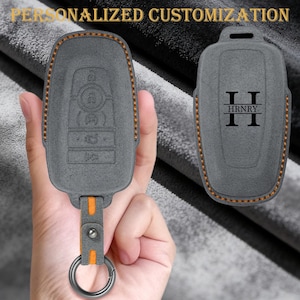 Peut inclure: Housse de clé grise avec coutures orange et une sangle orange assortie. La housse comporte un symbole de clé et d'autres boutons. La seconde housse porte les initiales "II" et le nom "HRNRY". Le texte "PERSONNALISATION PERSONNALISÉE" est en haut.